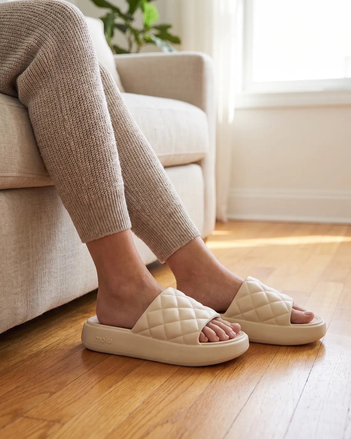 Pés calçando o chinelo molu Luxo bege, de tira matelassê, no piso de madeira da sala